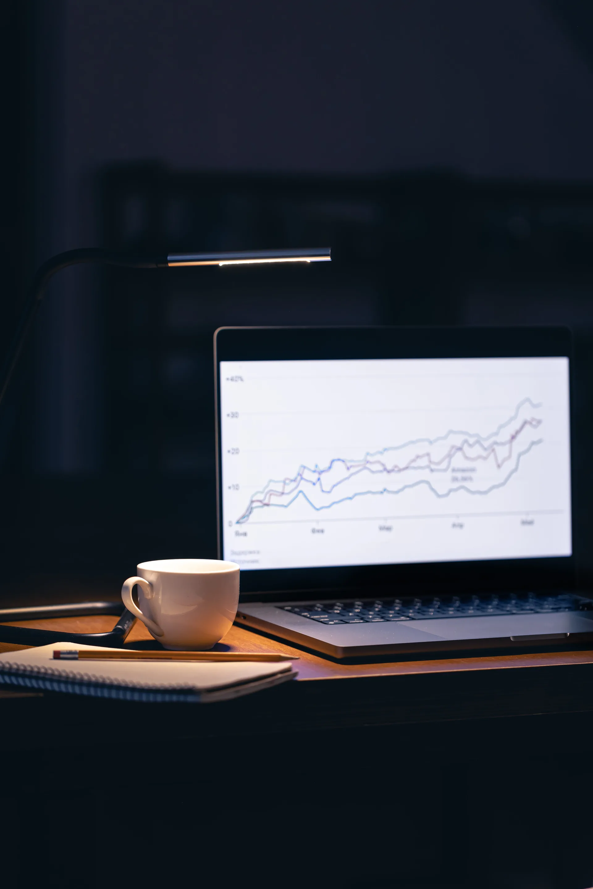 A laptop sat on a desk in a dark room, a mug of coffee is next to the laptop with a notebook and pen